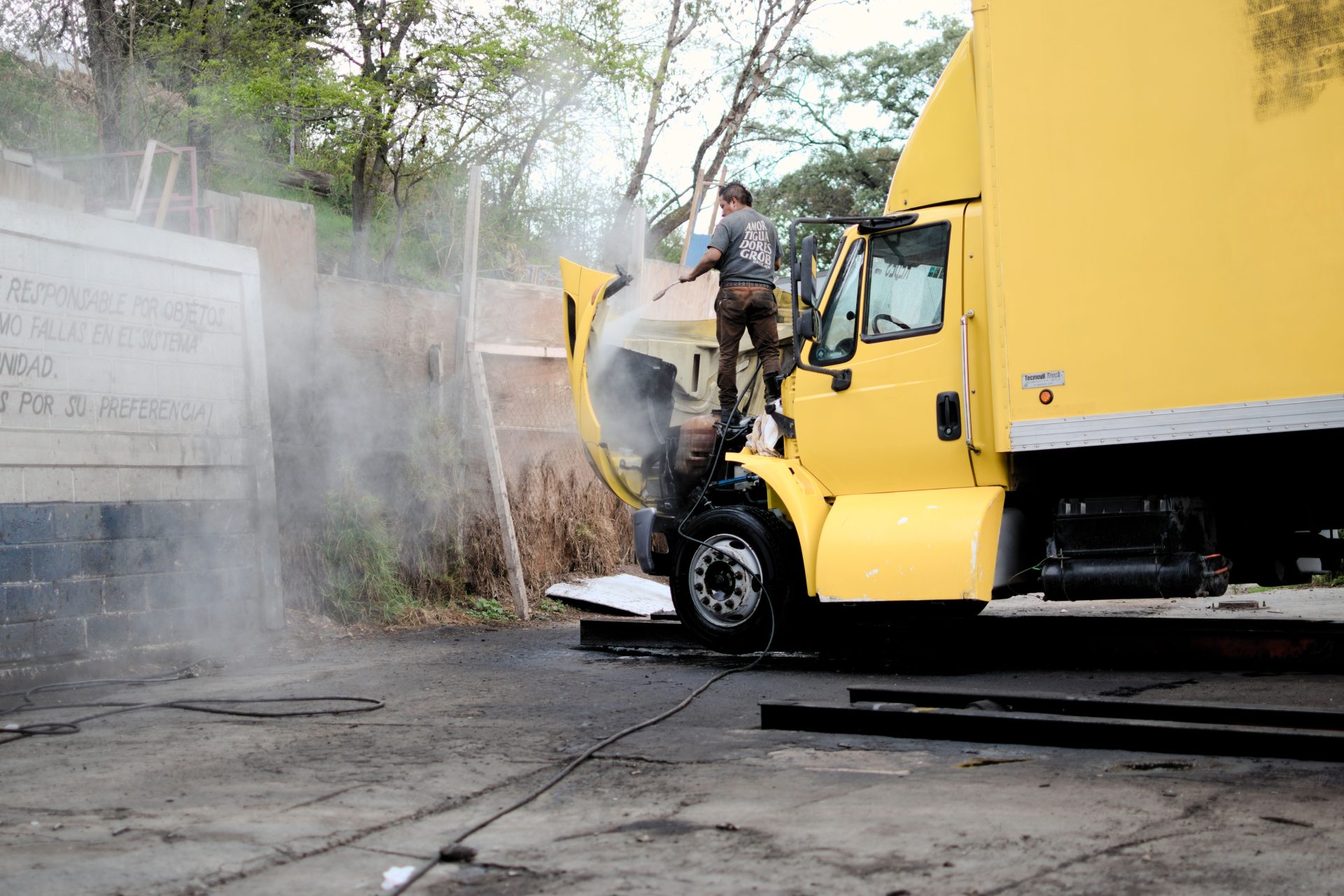 A mechanic inspects and repairs the engine of a yellow truck in an outdoor setting, surrounded by steam.