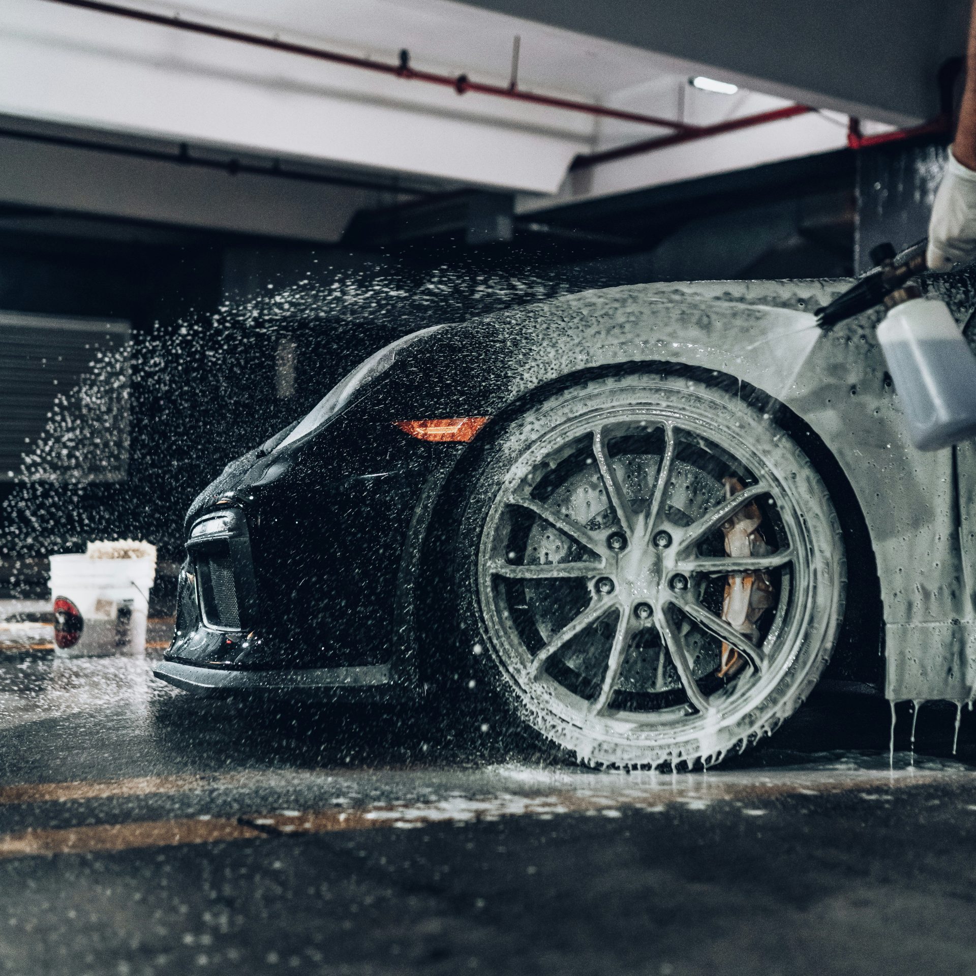 A person spraying soapy foam onto a black sports car in a garage